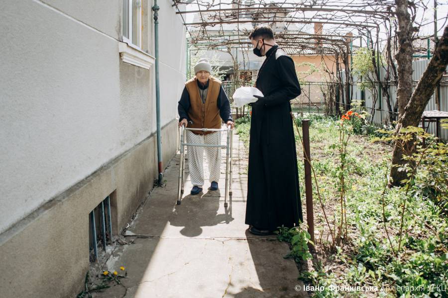 Eine alte Frau mit Gehhilfe in ihrem Garten mit einem Freiwilligen, der ihr ein Essenspaket bringt.