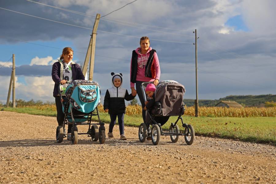 Zwei Frauen mit Kinderwagen und einem Kleinkind auf einer staubigen Schotterstraße.