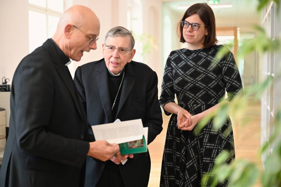 Im Bild stehen drei Personen in einem hellen Flur mit Holzboden zusammen. Links ist Prof. Thomas Schwartz zu sehen, bekleidet mit schwarzer Priesterkleidung und Brille. Er hält ein aufgeschlagenes Buch in den Händen und spricht darüber. In der Mitte steht Kardinal Kurt Koch, ebenfalls in schwarzer Priesterkleidung. Er hört aufmerksam zu und schaut interessiert auf das Buch. Rechts daneben steht Christiana Hägele. Im Vordergrund rechts sind einige grüne Pflanzen leicht verschwommen zu sehen.