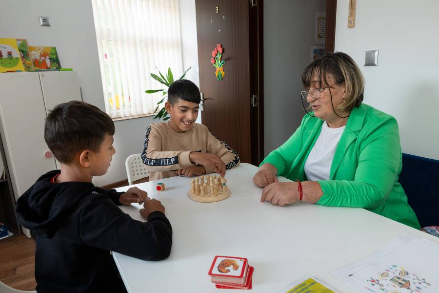 Zwei Kinder sitzen gemeinsam mit einer erwachsenen Frau am Tisch. Sie spielen ein Spiel, bei dem kleine Holzfiguren auf einem Spielbrett bewegt werden.