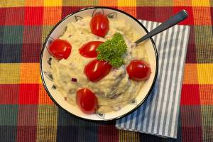 Eine Schüssel mit einem kleinen Löffel und einer gestreiften Serviette steht auf einem karierten Tischset, darin eine hellgelbe Auberginencreme mit kleinen Stückchen von roten Zwiebeln, obendrauf halbierte rote Cocktailtomaten und ein bisschen Petersilie.